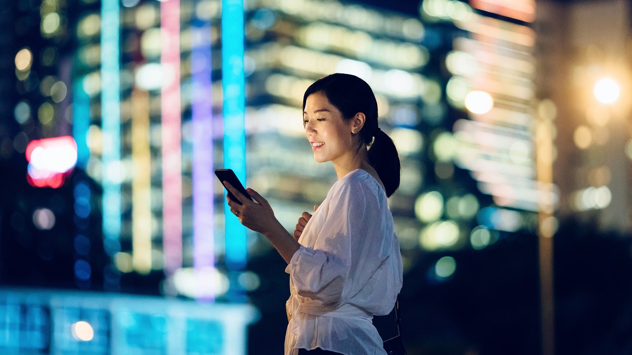 A girl is looking at her mobile phone against glowing city skyscrapers at night; image used for HSBC Taiwan i-invest for online trading page.