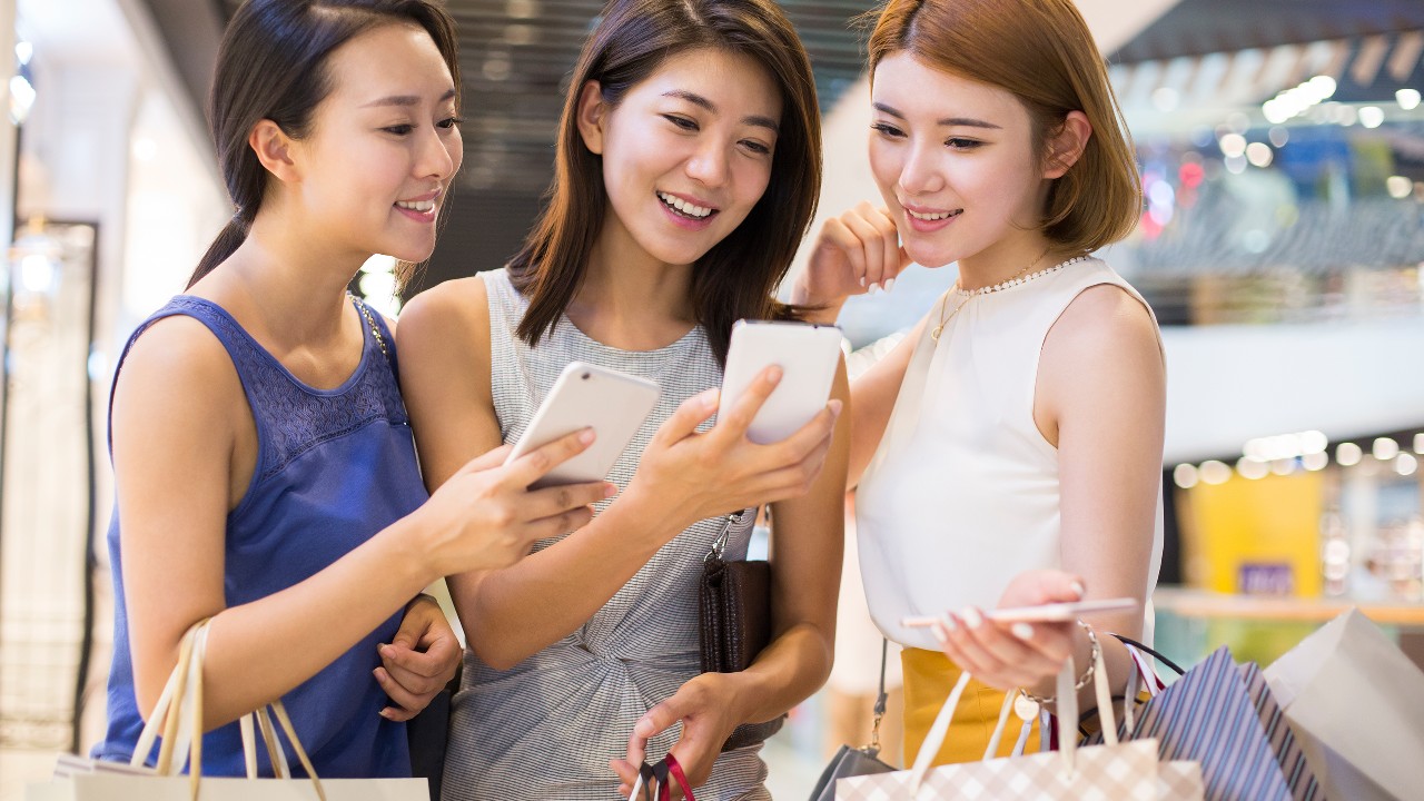 Three girls are looking at their mobile phone in a shopping mall; image used for HSBC Taiwan i-invest for online trading page.